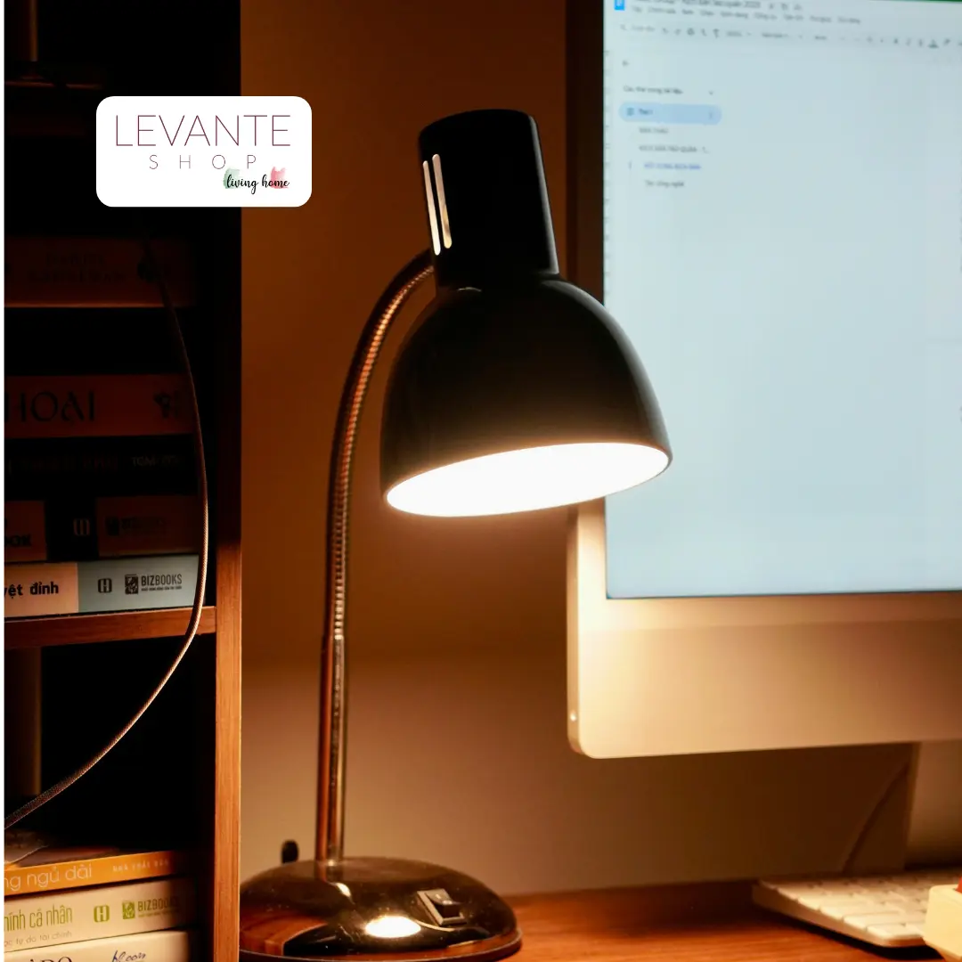Lampada da tavolo moderna accesa su scrivania accanto a monitor e libreria, perfetta per illuminare studio o angolo lavoro.